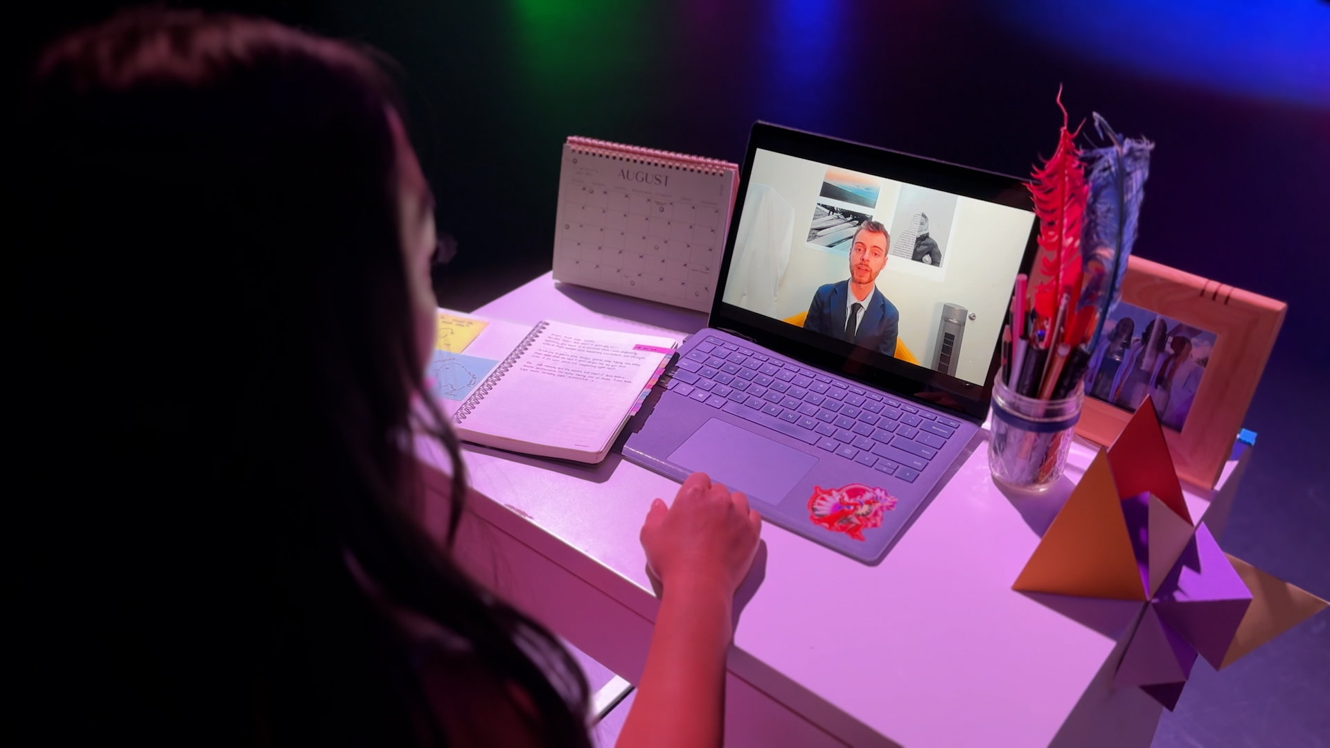 A person at a desk watches a suited man on a laptop video call, with a notebook, calendar, and props.