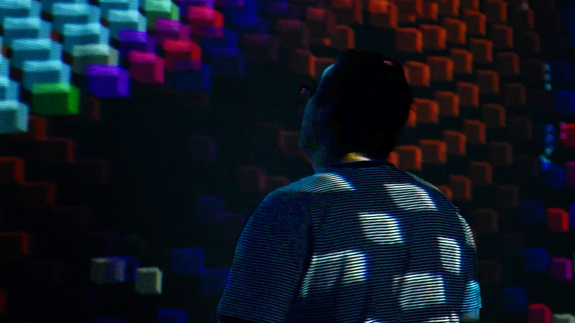 A person stands amid an immersive projection of glowing colorful pixel blocks in a dark room.