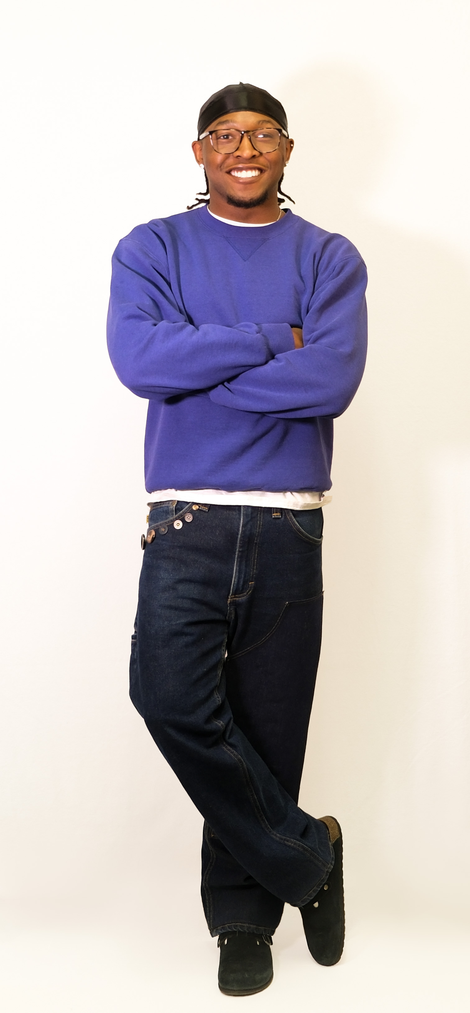 A smiling man poses against a white backdrop in a purple sweatshirt and dark baggy jeans.