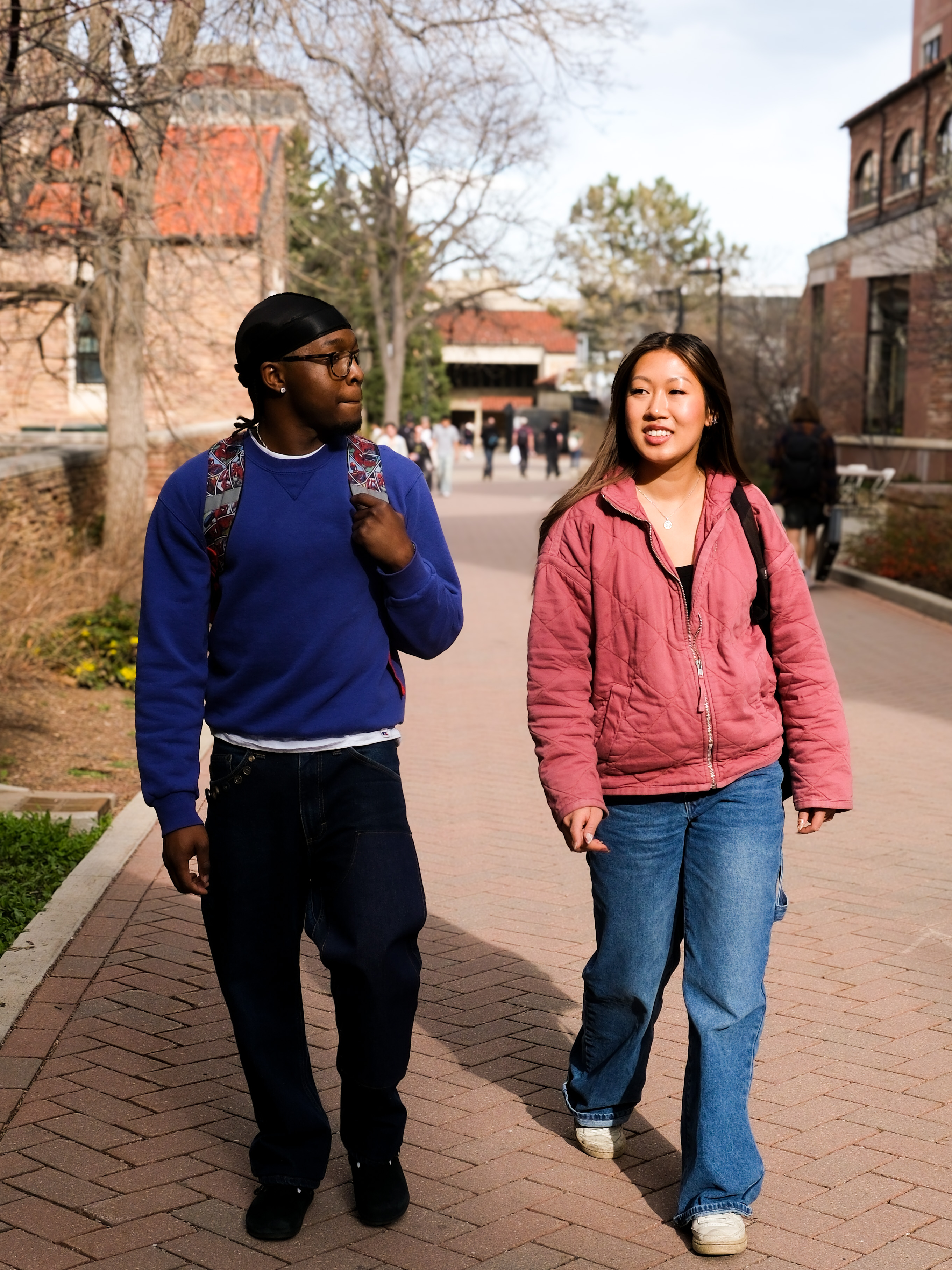Two students walk a campus path in upcycled clothing, a purple sweatshirt and a pink quilted jacket.