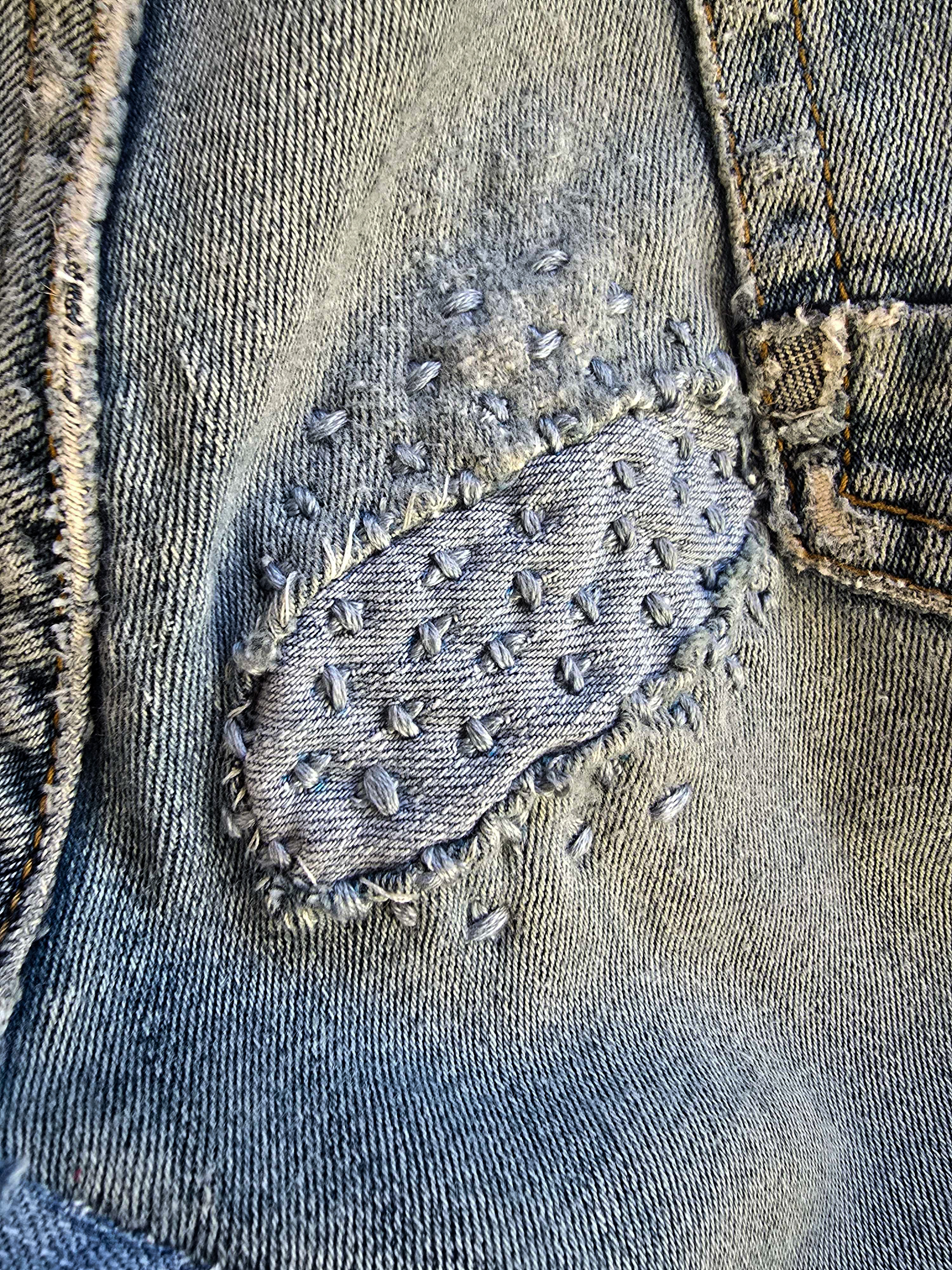Close-up of a hand-stitched denim patch with visible mending stitches on worn jeans.