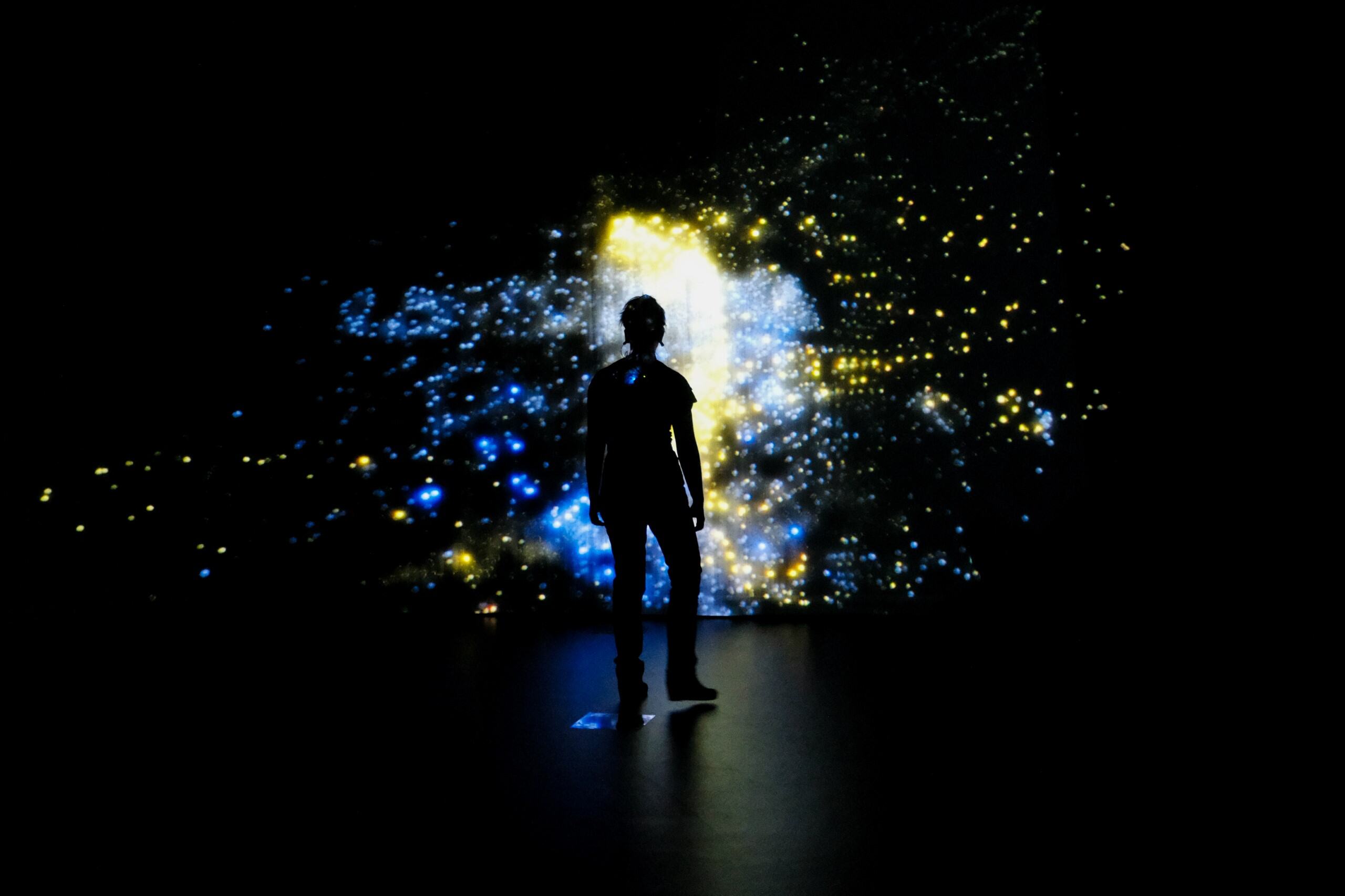 A silhouetted person stands before a glowing blue and gold particle projection in a dark installation.