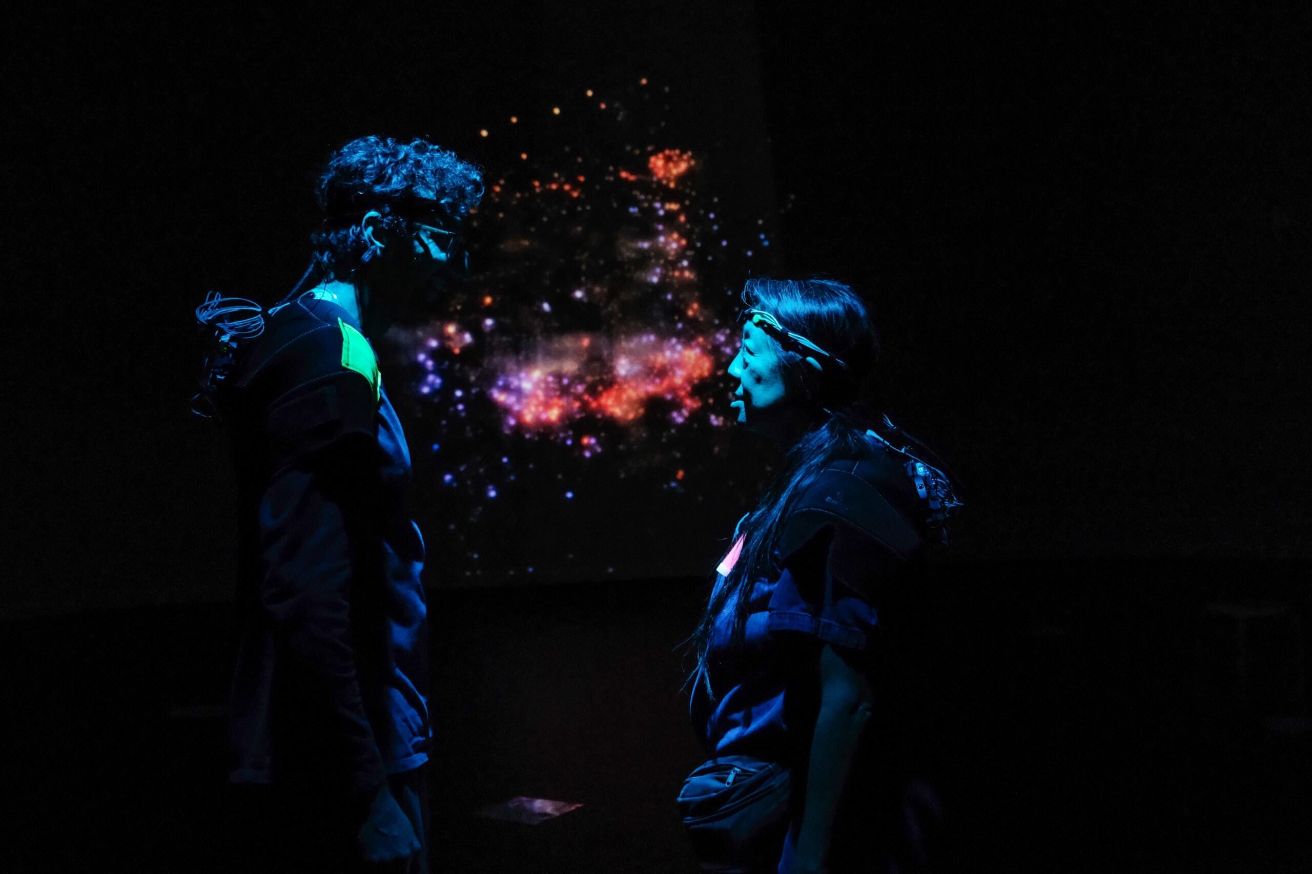 Two people wearing biometric headsets face each other before a glowing particle projection in the dark.