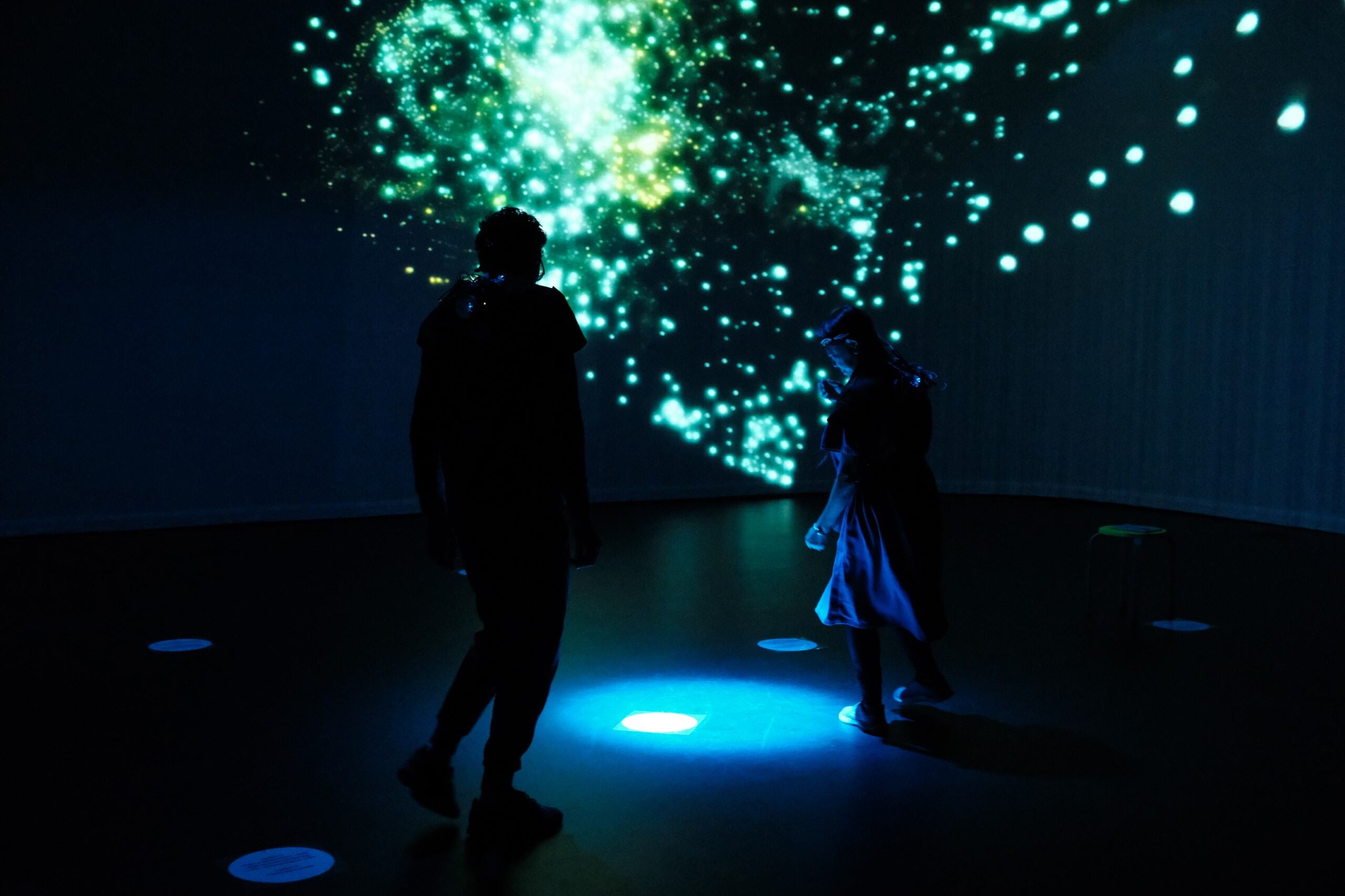 Two silhouetted people in a dark room with glowing green and blue particle projections and lit floor markers.