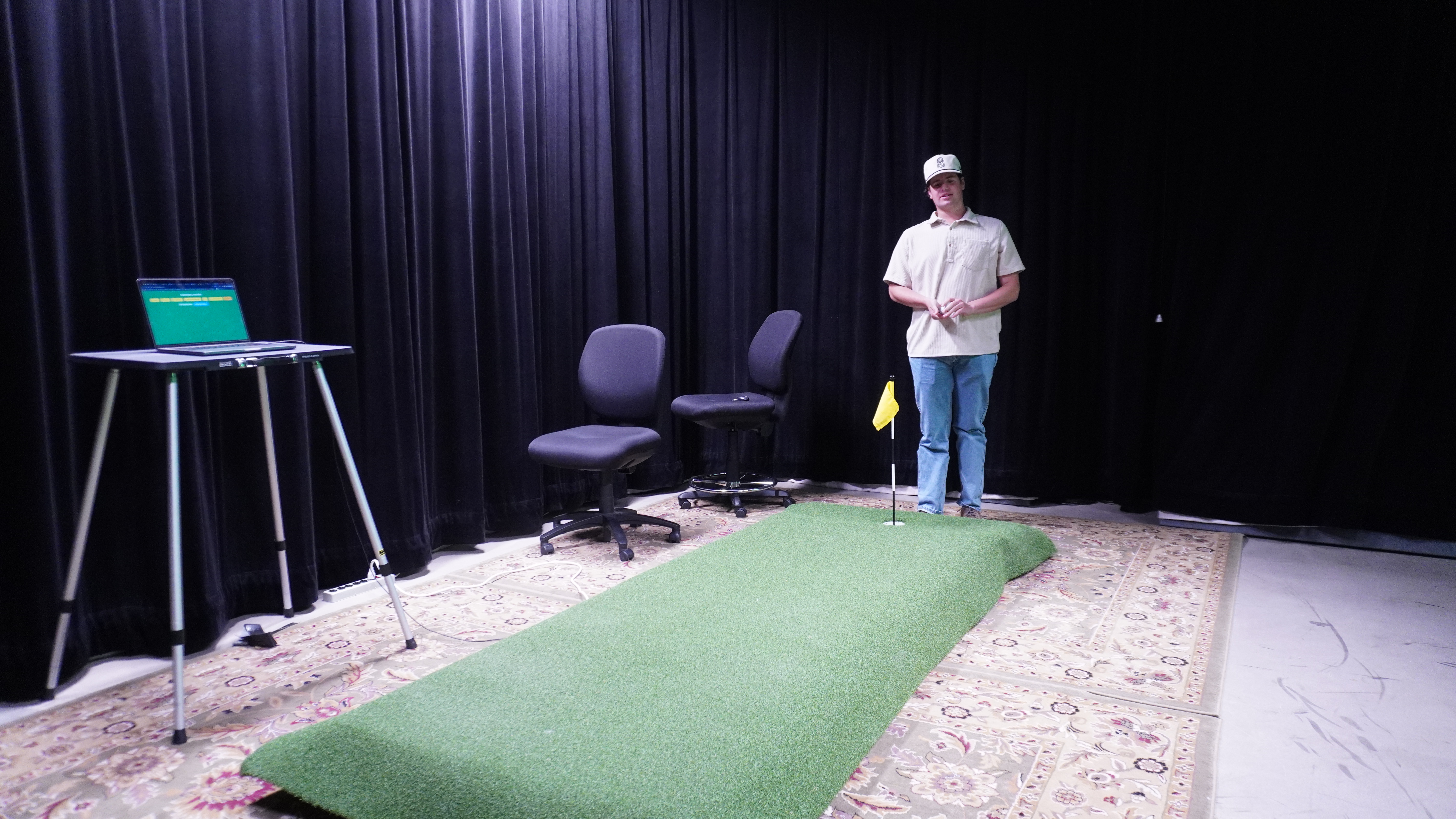 A student stands by a green putting mat with a flagged hole; a laptop on a stand sits nearby.