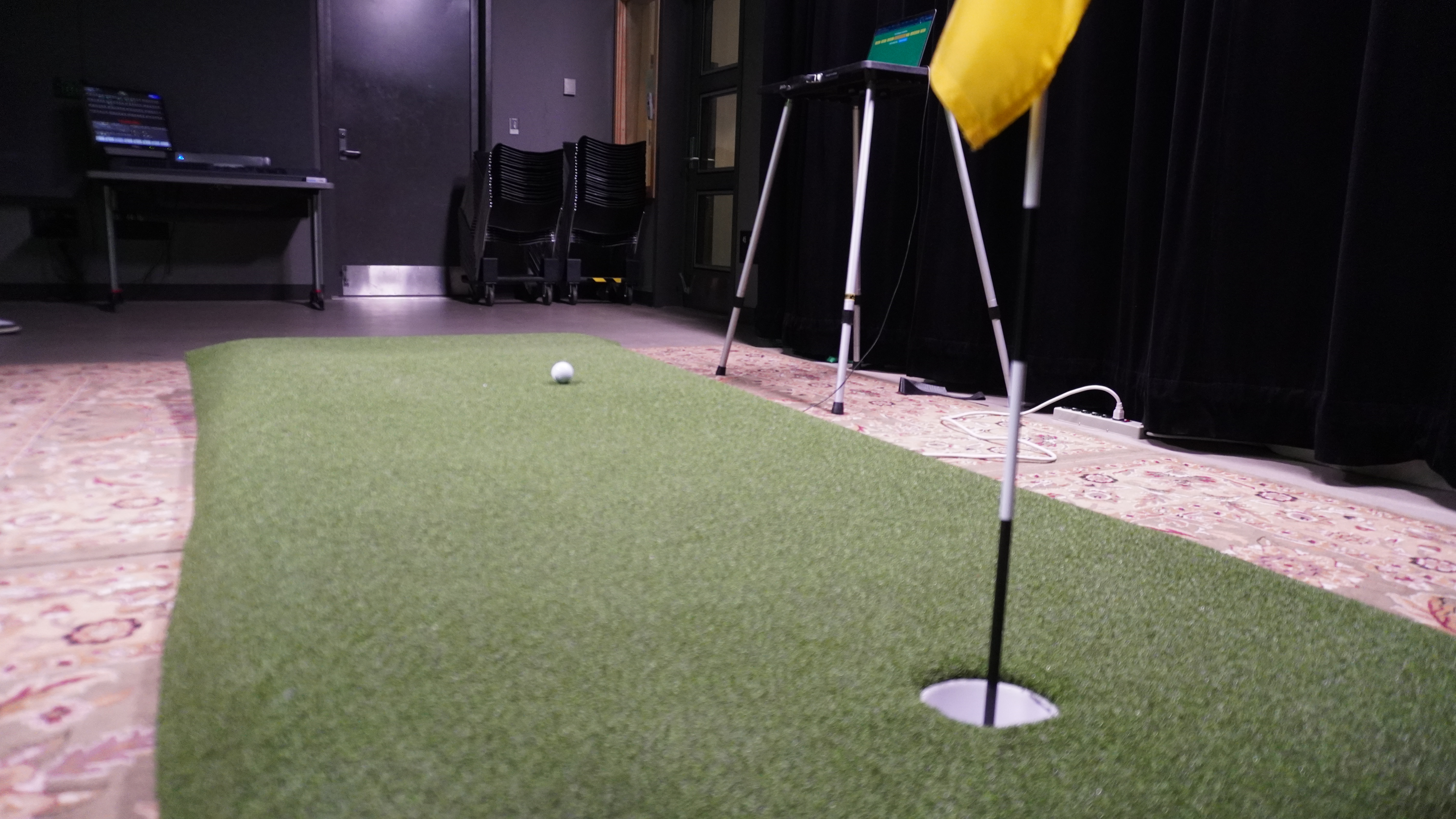 A green putting mat with a golf ball rolling toward a flagged hole, laptops on stands in the background.