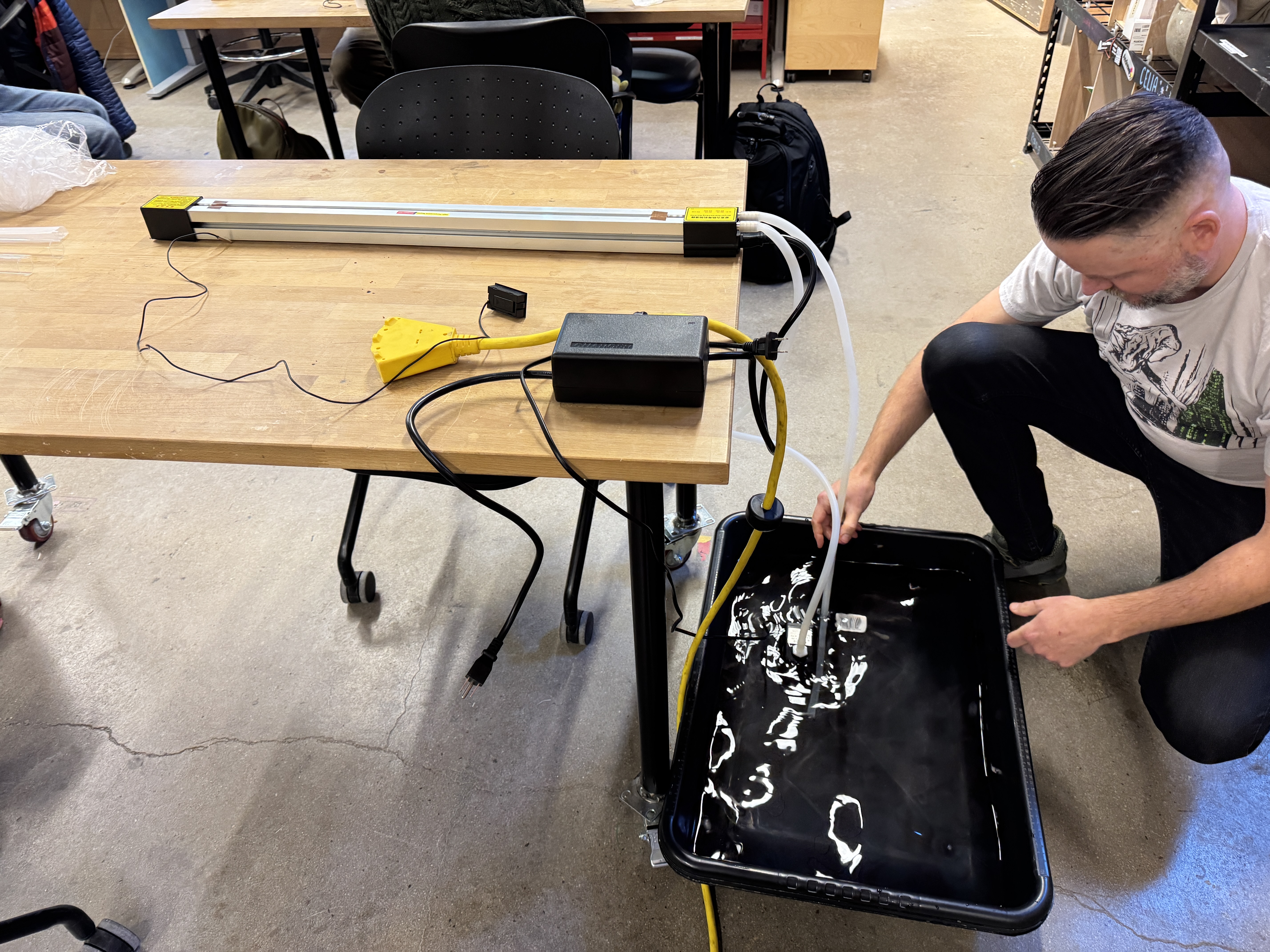 A man crouches in a workshop feeding tubing into a black tub, beside a long acrylic light fixture.