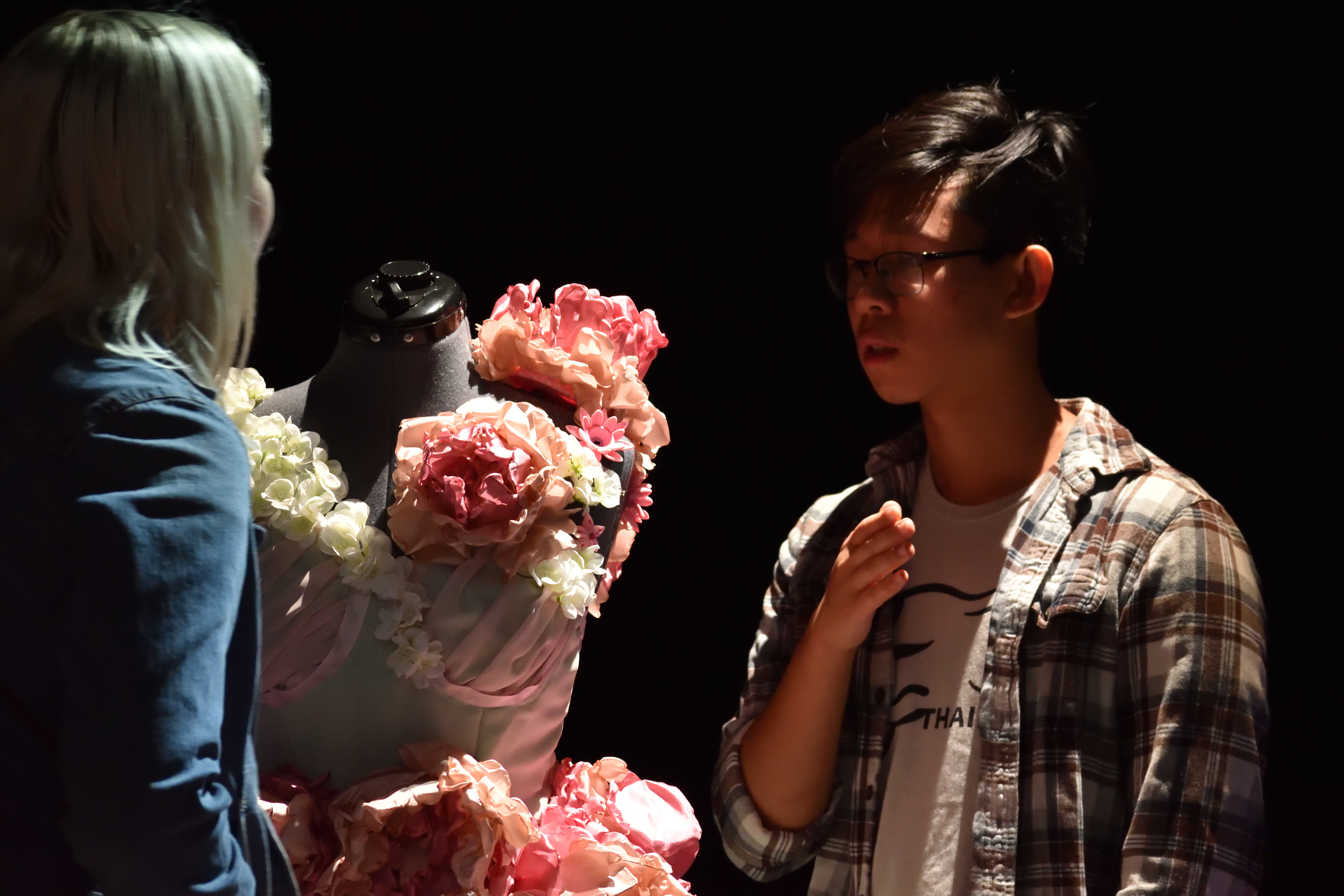 A bespectacled student gestures while discussing the floral gown installation with a visitor.