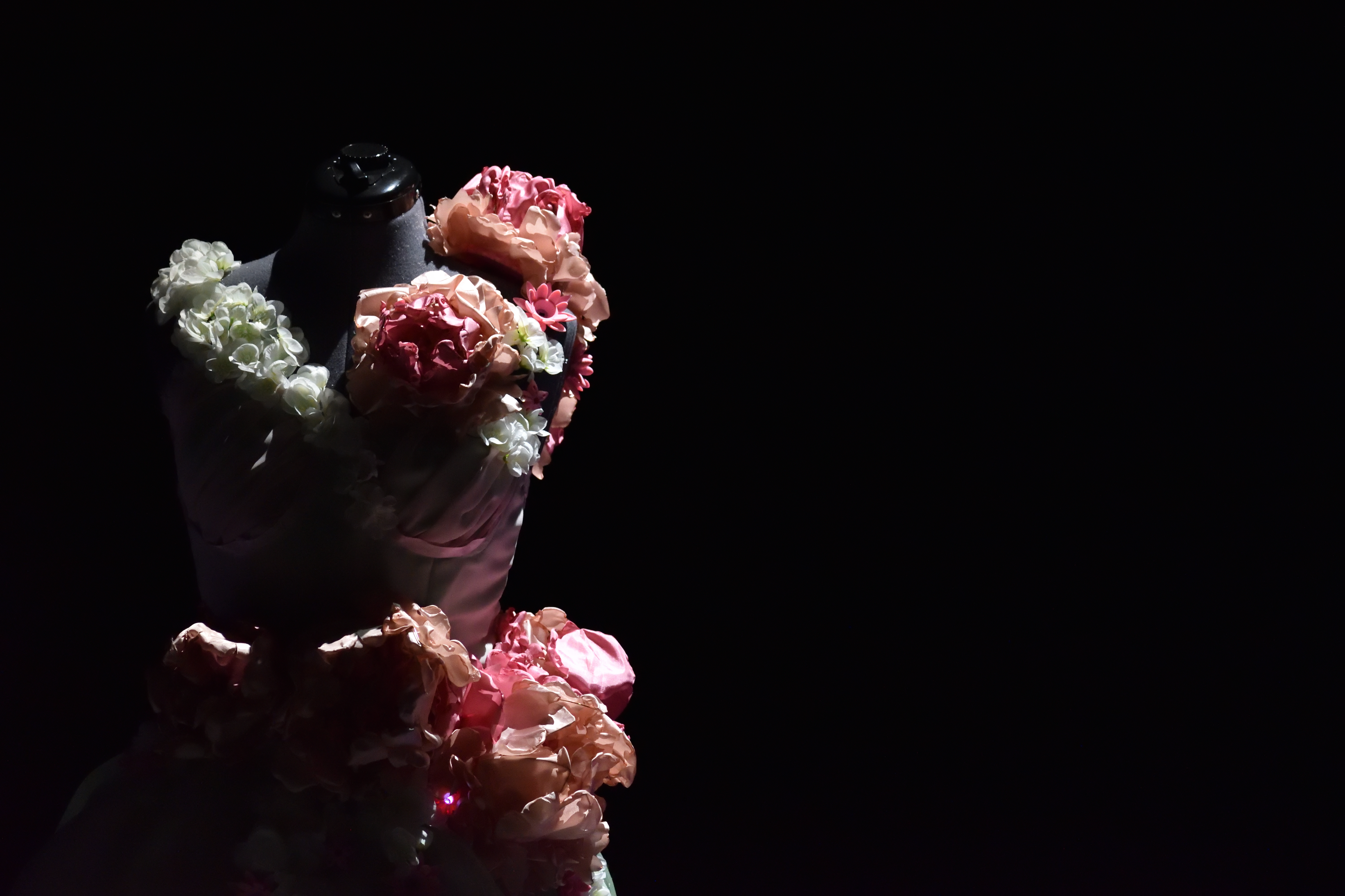 Floral-covered mannequin gown spotlit against a black background, flowers across the bodice.