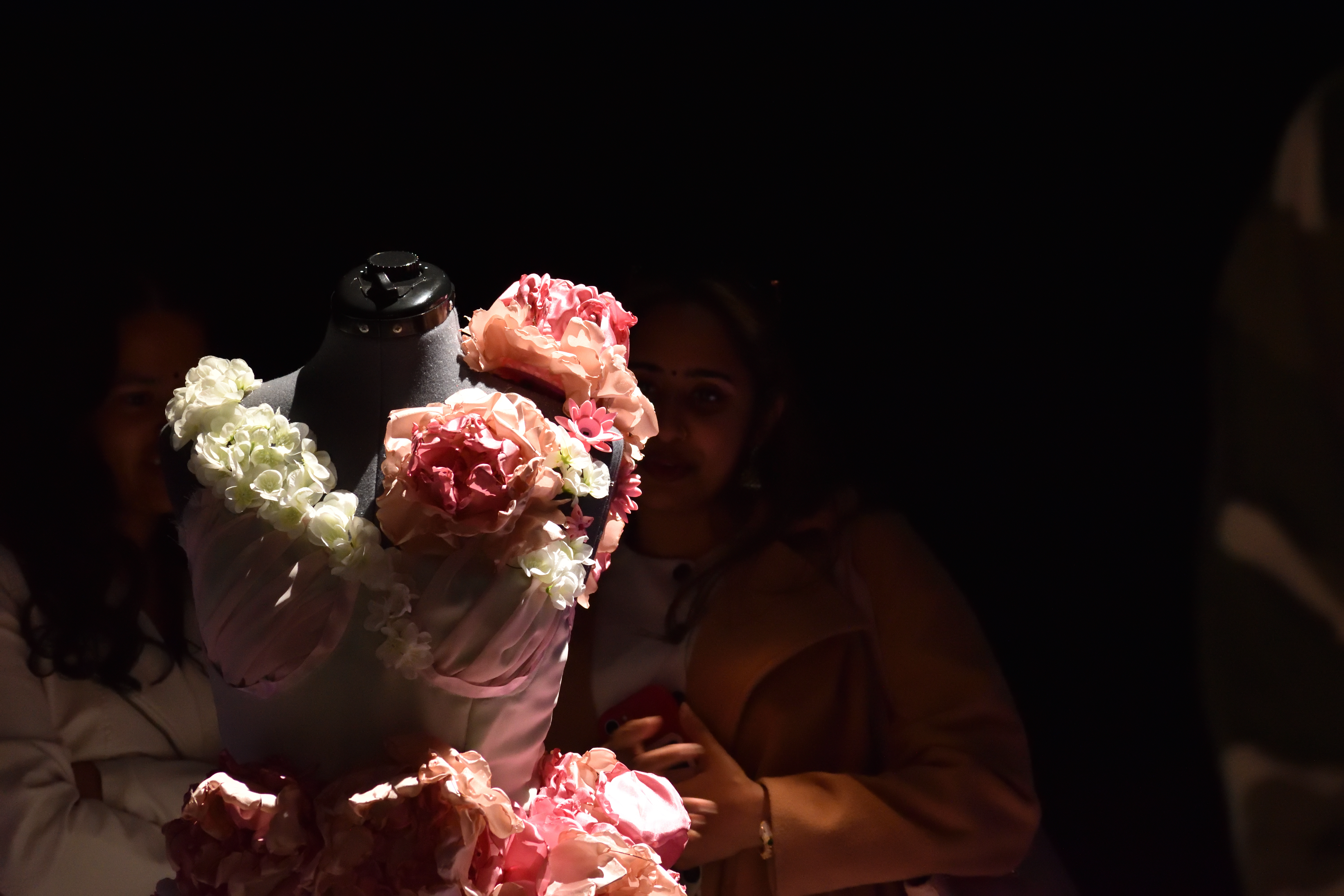 Visitors admire the flower-covered LED gown in a darkened gallery, one filming on her phone.
