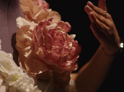 A hand reaches toward a large pink fabric peony on the flower-covered gown in low light.